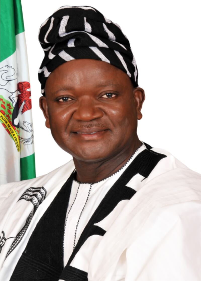 portrait of the Benue State Governor, on a white background with the Nigerian flag close to him, he is wearing a white and black striped cap